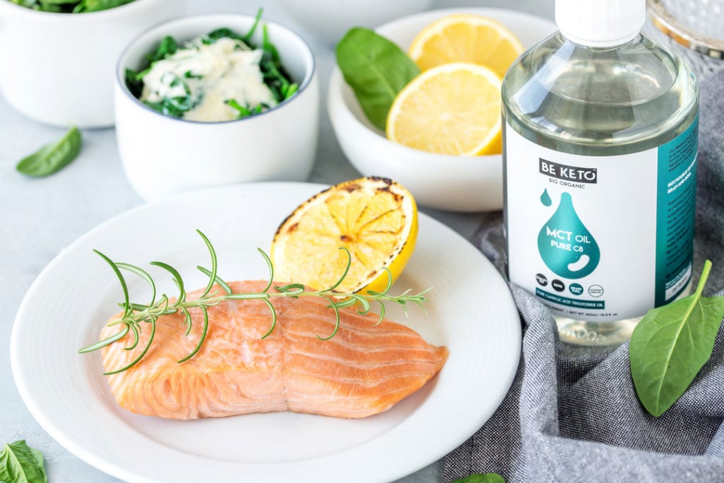 Keto Lachs mit Spinat: gebackenes Lachsfilet mit Rosmarin und gerösteter Zitrone auf einem weißen Teller, dahinter Rahmspinat, Zitronen und BeKeto MCT-Öl C8