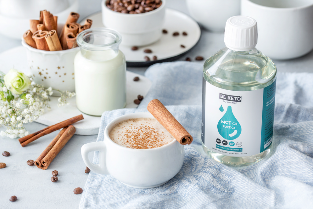 Keto Kaffee mit Zimt in einer weißen Tasse, daneben Zimtstangen, Mandelmilch und BeKeto MCT-Öl Pure C8