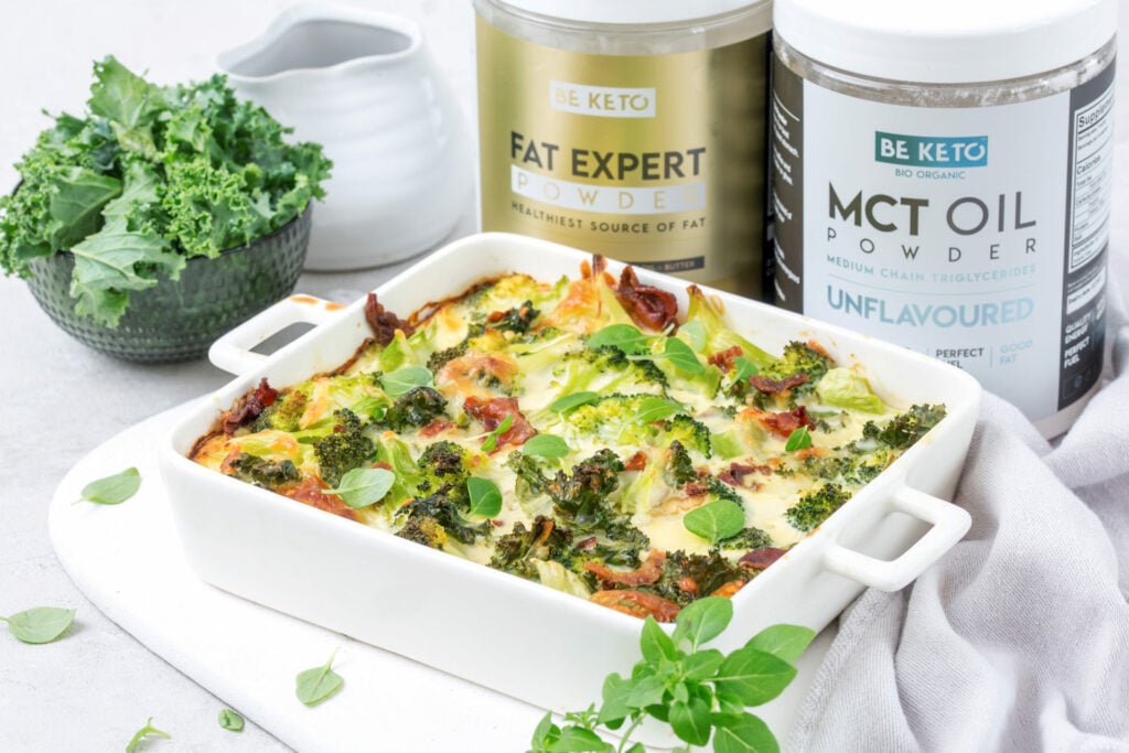 Keto Auflauf mit Brokkoli, Speck und Grünkohl in einer weißen Auflaufform, dahinter BeKeto Fat Expert Pulver und MCT-Öl-Pulver