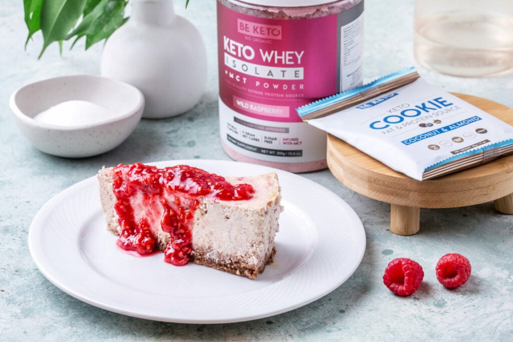 Stück Keto Himbeer-Käsekuchen mit Mandelboden und Himbeersoße, dahinter BeKeto Keto Whey Isolat Wild Raspberry und Keto Cookie Kokosnuss & Mandel