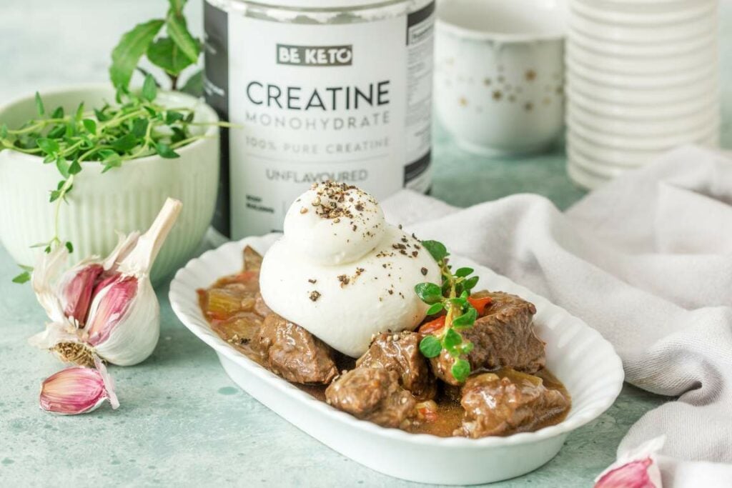Keto Gulasch aus Rindfleisch mit Burrata und frischen Kräutern in einer weißen Schale, dahinter BeKeto Creatine Monohydrate