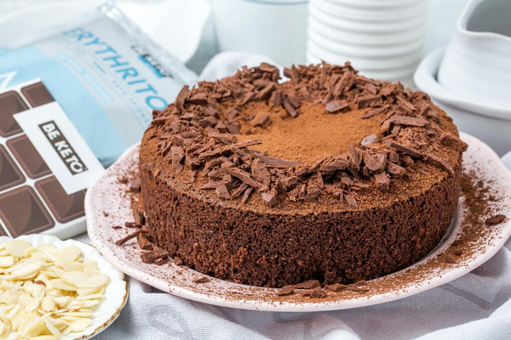 Keto Torte mit Schokolade, mit Kakao bestäubt und Schokoladenraspeln belegt, daneben BeKeto Erythritol, Keto-Schokolade Classic Dark und Mandelblättchen