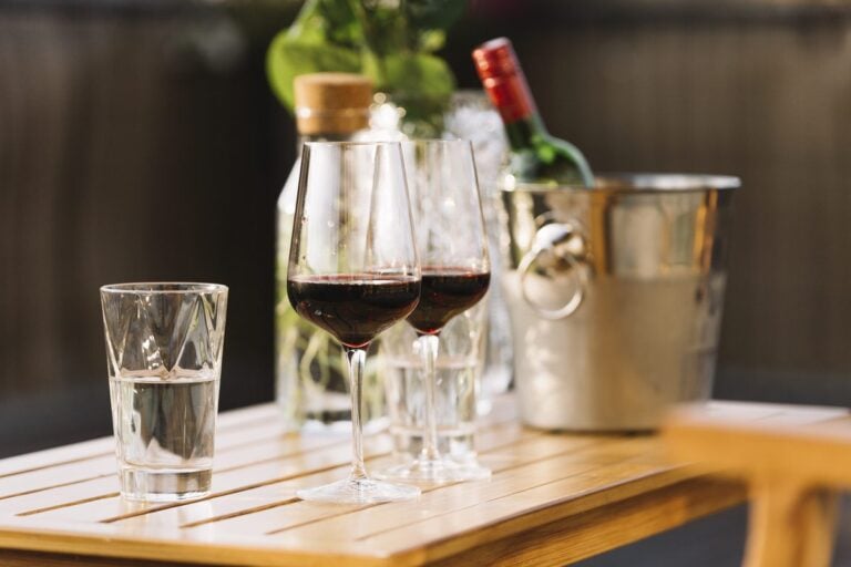 Zwei Gläser Rotwein und ein Glas Wasser auf einem Holztisch, dahinter ein Weinkühler