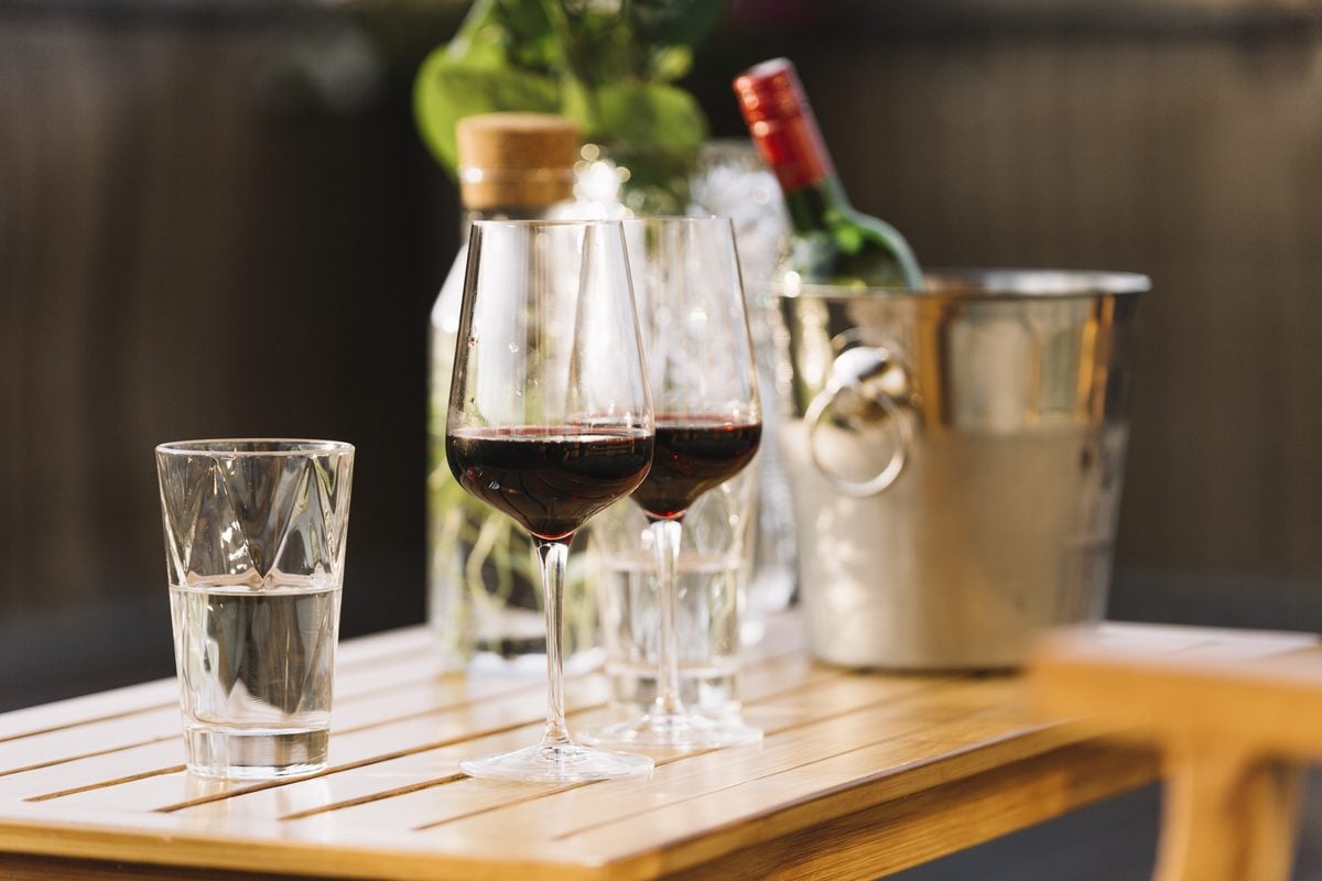 Alkohol bei Keto: Was erlaubt ist und was er bremst 1 Zwei Gläser Rotwein und ein Glas Wasser auf einem Holztisch, dahinter ein Weinkühler