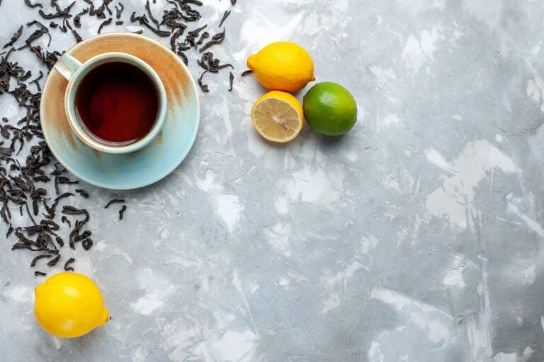 Tasse schwarzer Tee auf einer Untertasse, daneben lose Teeblätter, Zitronen und eine Limette auf hellem Grund