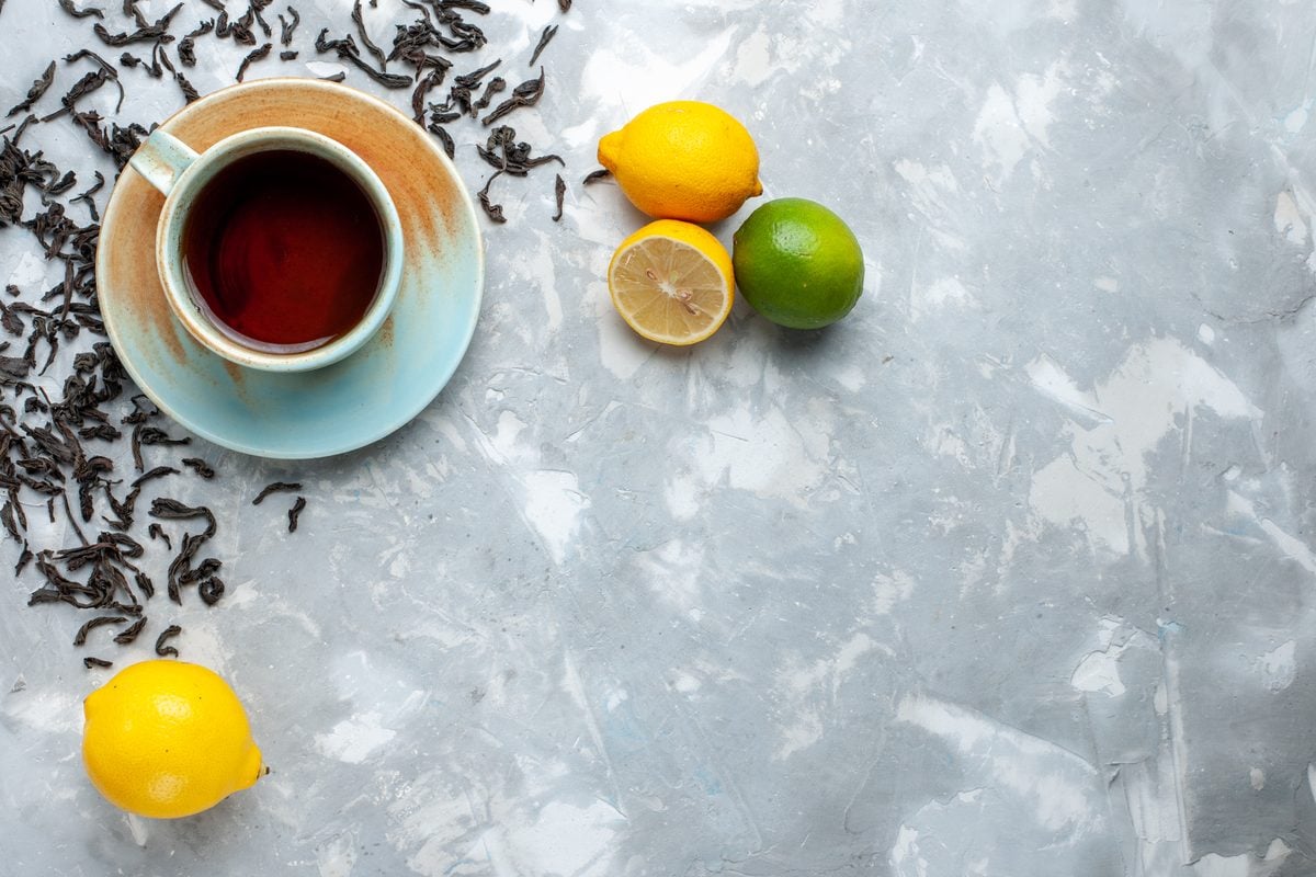 Keto Getränke: Was man trinken darf 1 Tasse schwarzer Tee auf einer Untertasse, daneben lose Teeblätter, Zitronen und eine Limette auf hellem Grund