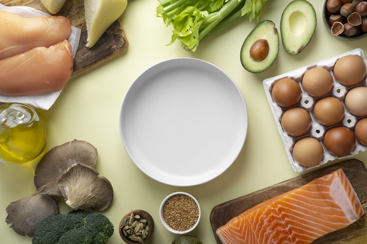 Keto Makros: die richtige Verteilung von Fett, Eiweiß und Kohlenhydraten 1 Hähnchenbrust, Lachsfilet, Eier, Avocado, Olivenöl, Pilze, Brokkoli und Nüsse rund um einen leeren Teller