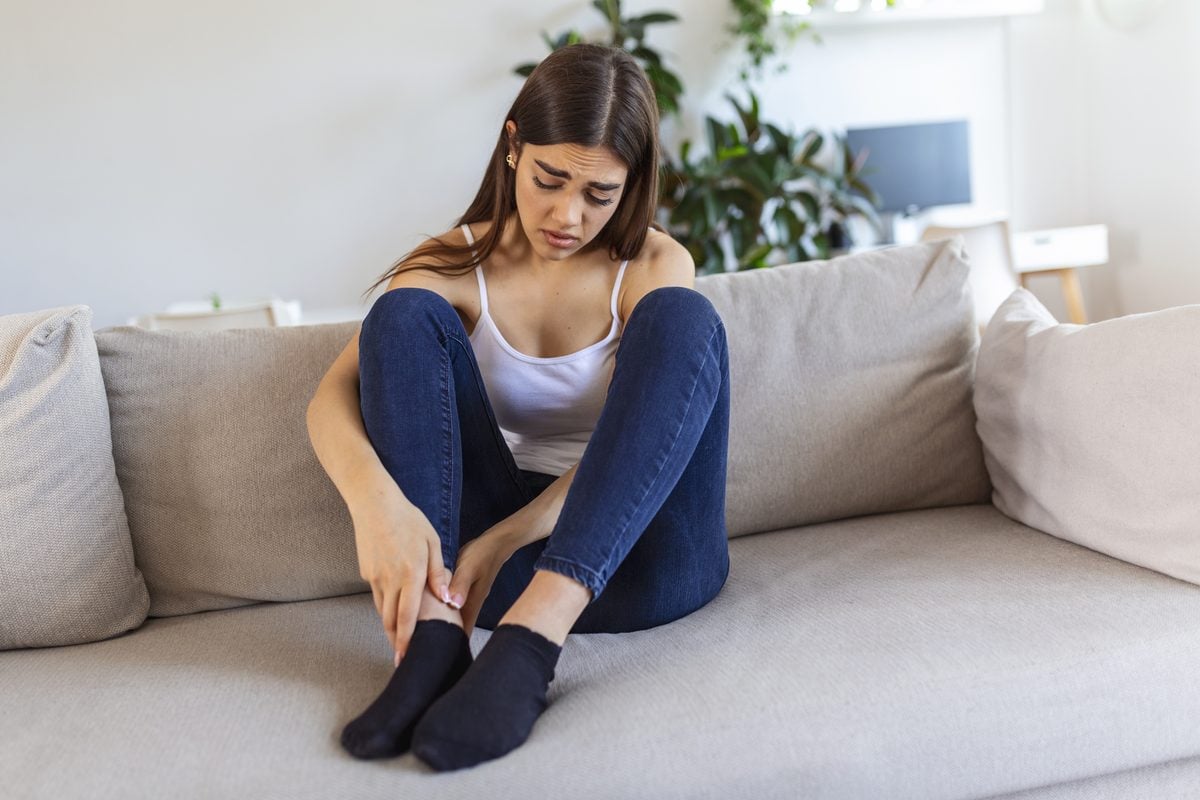 Keto Nebenwirkungen: Was vorübergehend ist und was Aufmerksamkeit braucht 1 Junge Frau sitzt auf dem Sofa und massiert ihren schmerzenden Unterschenkel