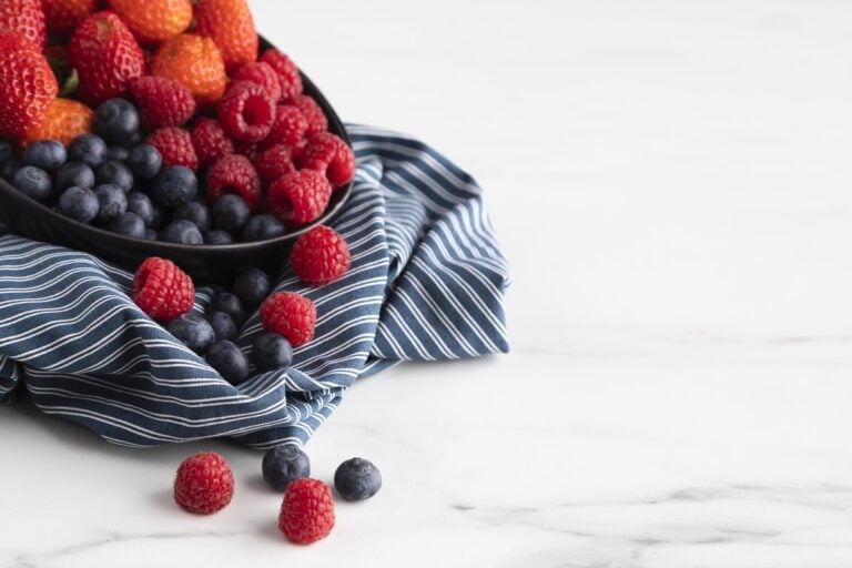 Schale mit Erdbeeren, Himbeeren und Heidelbeeren auf einem gestreiften Tuch, daneben einzelne Beeren auf Marmor