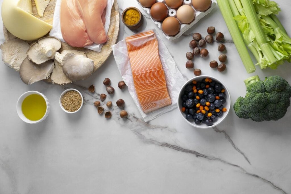 Lachsfilet, Hähnchenbrust, Eier, Käse, Pilze, Nüsse, Olivenöl, Staudensellerie, Brokkoli und Beeren auf hellem Marmor