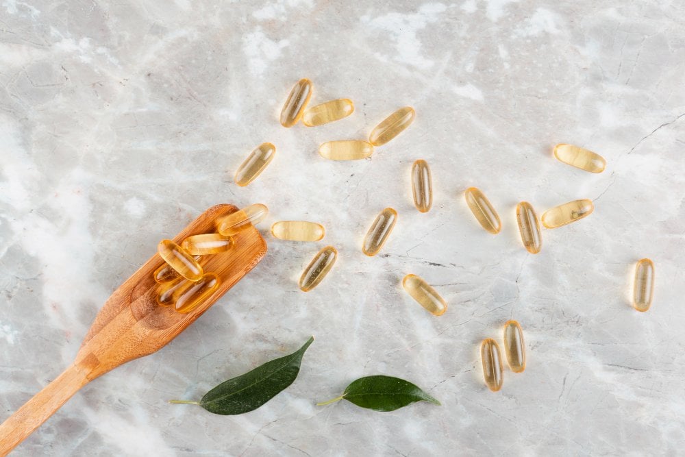 Omega-3-Kapseln auf hellem Marmor, ein Teil davon auf einem Holzlöffel, daneben zwei grüne Blätter